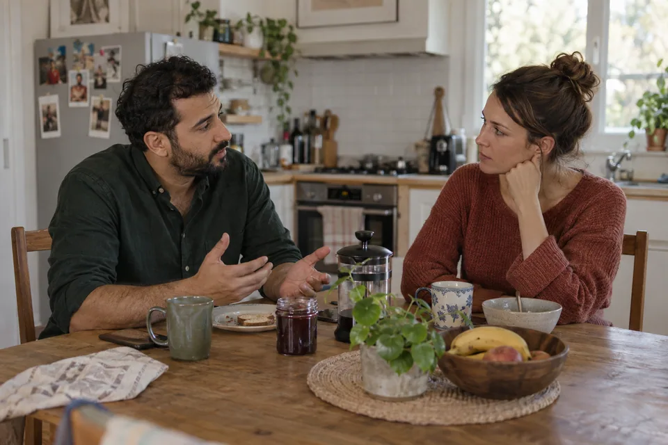 Two partners sit at a kitchen table and listen to each other.